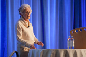 Jean Hamilton stands behind a table with a white tablecloth on it. she is wearing a white sweater and has short white hair and glasses.