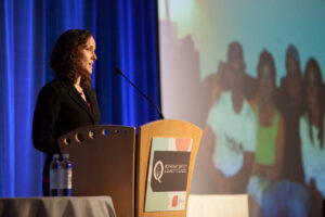 Sarah Chapdelaine stands behind a podium on stage. To her right is a large photo on a screen that is out of focus. Sarah is wearing a black blazer and has brown curly hair.