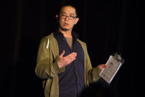 David Ng presents on stage while holding a clipboard. He is mid-sentence with his hand gesturing beside him. He is wearing a green hoodie, blue button down shirt, glasses and has short dark hair.