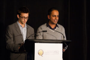 Naheed Dosani and Stephen Pomedli stand beside one another on stage behind a podium presenting.