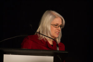 Judy Pryce stands behind a podium on stage. She is wearing a red jacket, glasses and has shoulder-length silver hair.