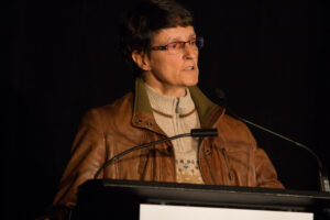 Lise Mathieu stands on stage behind a podium presenting. She is wearing a tan jacket with a tan sweater underneath, glasses and has short dark hair.